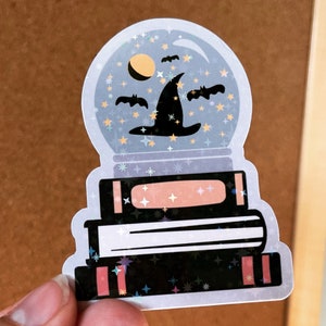 May include: A glass snow globe with a witch's hat and bats inside, sitting on top of a stack of three books. The snow globe and books are decorated with glitter.