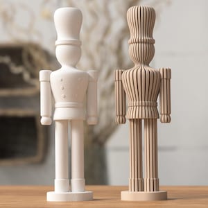 Könnte beinhalten: Zwei Holz-Nussknacker-Figuren. Eine ist weiß lackiert, die andere ist aus Naturholz mit Streifenmuster. Beide haben ein soldatenähnliches Aussehen mit hohen Hüten und detaillierten Uniformen.