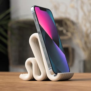 Könnte beinhalten: Ein cremefarbener Telefonständer mit einem einzigartigen, wellenförmigen Design. Er hält ein Smartphone mit einem farbenfrohen Bildschirm. Der Ständer besteht aus einem glatten Material und steht auf einer Holzoberfläche.