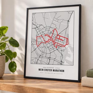 Op de afbeelding: Een ingelijste kaartprint met een grijs en wit stratenplan van een stad en een gemarkeerde rode marathonroute. De tekst luidt Anna Schneider 412345, Mein Erster Marathon en 42,2 km. De print zit in een dunne zwarte lijst met witte rand.