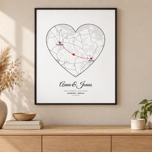Puede incluir: Una lámina de arte enmarcada con un mapa en forma de corazón con una línea roja que conecta Hamburgo y Berlín, marcada por pequeños corazones rojos. Debajo del mapa, el texto dice Anna & Jonas, ZWEI STÄDTE, EIN HERZ, HAMBURG - BERLIN, 14.02.2018.