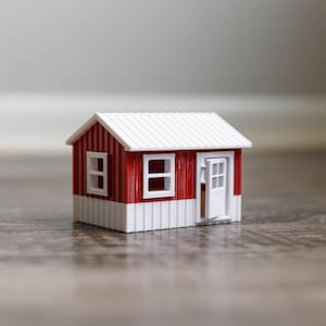 Puede incluir: Una pequeña casa de juguete roja y blanca con una puerta blanca y dos ventanas. La casa está sentada en un suelo de madera.