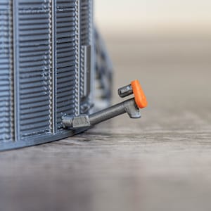 May include: A close-up of a grey metal model train with an orange safety light on the side.