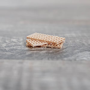 May include: A small stack of miniature wooden pallets. Each pallet has a rectangular shape and a grid-like pattern. The pallets are light brown in colour and appear to be made of wood. The background is a blurred, grey wooden surface.