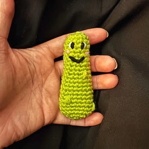 May include: A handmade, green crocheted figure with a smiling face, held in a hand. The figure has black eyes, a black smile, and pink cheeks. The background is a dark fabric.