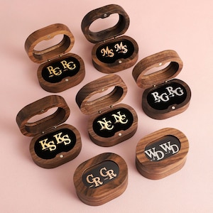 May include: Seven oval-shaped dark wood boxes with hinged lids and clear windows. Each box contains a pair of metallic cufflinks featuring various monogrammed initials in gold or silver tones, displayed against a black velvet interior lining.