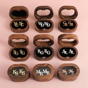 Könnte beinhalten: Neun ovale Schatullen aus dunklem Holz mit Klappdeckeln, die jeweils ein Paar metallische Manschettenknöpfe enthalten. Die Manschettenknöpfe zeigen verschiedene zweistellige Monogramm-Kombinationen in Gold- oder Silbertönen auf schwarzem Samt.