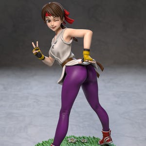 Op de afbeelding: Figuur van een vrouw in een witte tanktop, paarse legging en rode laarzen. Ze knipoogt en geeft het vredesteken. De figuur staat op een groene basis met witte bloemen en een stenen pad.