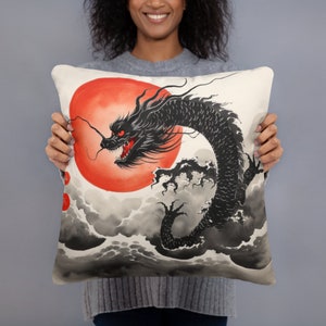 Könnte beinhalten: Quadratisches Kissen mit einem schwarzen Drachen mit roten Augen und offenem Maul vor einer großen roten Sonne. Der Drache ist vor einem Hintergrund aus grauen Wolken und Wellen platziert. Das Kissen hat einen cremefarbenen Hintergrund.