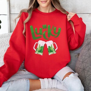 Può includere: Felpa rossa con una grafica verde "Lucky" e due boccali di birra verdi che brindano.