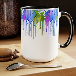 May include: White ceramic mug with a black rim and handle. The mug features a colorful abstract design of blue, green, and pink paint dripping down the front.