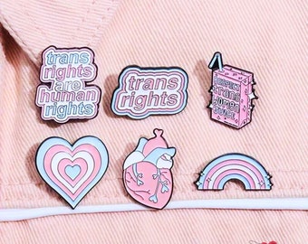 May include: A collection of enamel pins with LGBTQ+ themes. Designs include a heart, a rainbow, and text that reads "trans rights are human rights", "trans rights", and "Respect trans women juice". The pins are in shades of pink, blue, and white.