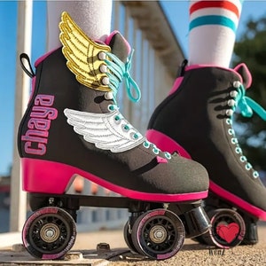 May include: Black roller skates with pink accents, wheels, and heels. The skates feature teal laces and the word "Chaya" on the side. Gold and white wing designs adorn the sides of the skates. The wheels are black with pink detailing.
