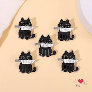 May include: Five black cat enamel pins, each holding a knife in its mouth. The knives have text that reads "THEY/THEM", "HE/HIM", and "SHE/HER". The pins are on a light beige background.