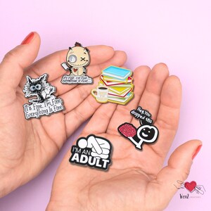 May include: A collection of enamel pins with humorous phrases and illustrations. Designs include a cat, a voodoo doll, a stack of books with a coffee cup, a brain, and a person lying down. Each pin has a black border and colorful details.