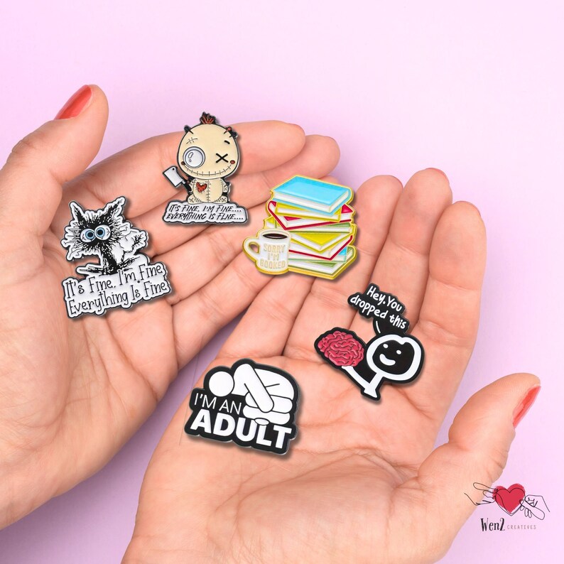 May include: A collection of enamel pins with humorous phrases and illustrations. Designs include a cat, a voodoo doll, a stack of books with a coffee cup, a brain, and a person lying down. Each pin has a black border and colorful details.