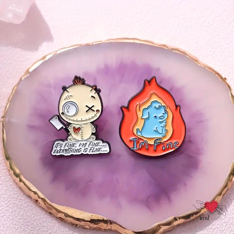 May include: Two enamel pins on a purple and gold-edged surface. One pin features a voodoo doll with a cleaver and the text "IT'S FINE, I'M FINE... EVERYTHING IS FINE...". The other pin shows a blue dog in flames with the text "I'M FINE".