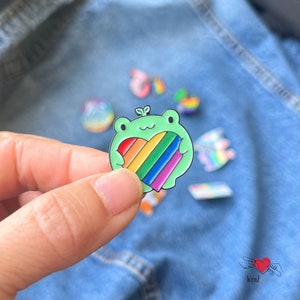 May include: A green frog pin holding a rainbow heart. The pin has a glossy finish and is set against a denim background. Other pins with rainbow designs are visible in the background.