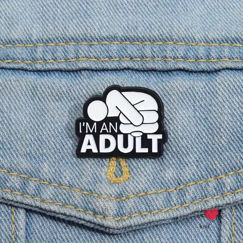 May include: A black and white enamel pin with the text "I'M AN ADULT" and a graphic of a person lying down. The pin is attached to a light blue denim jacket pocket.