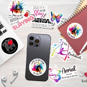 May include: A collection of colorful stickers with aerial and pole dancing themes. The stickers feature phrases like "Eat, Slay, Aerial, Repeat" and "Life is Better in the Air." The stickers are on a white surface with a laptop, phone, and notebook.