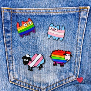May include: Four colorful enamel pins on a denim background. The pins feature a rainbow cat, a cat with the transgender flag, a sheep with the transgender flag, and a rainbow sheep. The pins are small and detailed.