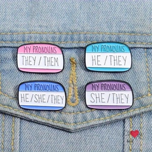 May include: Four enamel pins with the text "MY PRONOUNS" and different pronoun sets: "THEY/THEM", "HE/THEY", "HE/SHE/THEY", and "SHE/THEY". Each pin has a white background with a colored top and bottom border, set against a denim background.