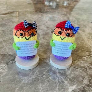 May include: Two handmade crochet figures with rainbow-colored bodies, wearing glasses and bows. Each figure holds a sign with the text: "Embracing one's authentic self is a powerful journey toward self-discovery and happiness!" They sit on wooden bases.