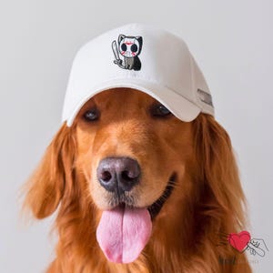 May include: A Golden Retriever wearing a white baseball cap with a black and red cat design. The cat is holding a knife and wearing a hockey mask. The dog has a pink tongue and is looking at the camera.