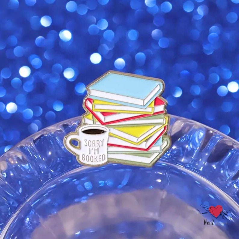 May include: A colorful enamel pin featuring a stack of books in blue, red, and yellow. A white mug with the text "Sorry I'm Booked" sits in front of the books. The pin has a gold outline and is set against a blue background.