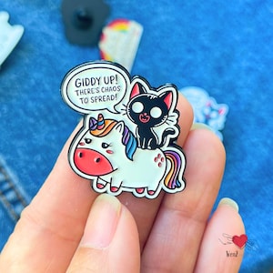 May include: A colorful enamel pin featuring a white unicorn with a rainbow mane and tail, ridden by a black cat. The pin includes a speech bubble with the text "Giddy Up! There's Chaos to Spread!"
