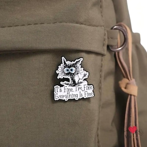 May include: A black and white enamel pin featuring a cartoon cat with wide blue eyes. The pin has the text "It's Fine. I'm Fine. Everything Is Fine" in a white rectangular border. The pin is attached to a green backpack.