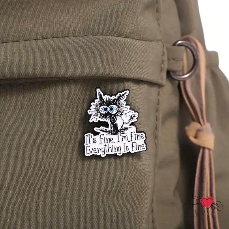 May include: A black and white enamel pin featuring a cartoon cat with wide blue eyes. The pin has the text "It's Fine. I'm Fine. Everything Is Fine" in a white rectangular border. The pin is attached to a green backpack.