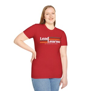 以下が含まれることがあります： 「Lead Learner」の白文字と茶色、オレンジ、グレーの横縞模様が入った赤いTシャツ。テキスト「CC Essentials Tutor」が下にあります。Tシャツは人が着ています。