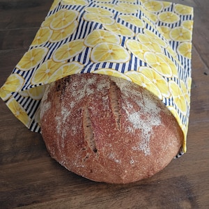 May include: A round, crusty loaf of brown bread partially tucked inside a fabric bag. The bag features a pattern of yellow lemon slices set against a background of thin, vertical navy blue and white stripes, creating a bright, contrasting look.
