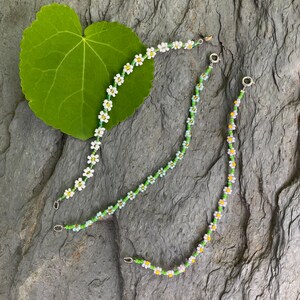 Pode incluir: Três pulseiras de contas com contas brancas em forma de margarida e detalhes verdes. As pulseiras estão sobre uma superfície cinzenta texturizada.