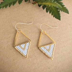 Peut inclure: Une paire de boucles d'oreilles géométriques dorées en forme de triangle. La partie inférieure de chaque boucle d'oreille est remplie de perles blanches et dorées selon un motif en chevrons. Les boucles d'oreilles sont présentées sur un fond neutre avec de la verdure et une fleur blanche.