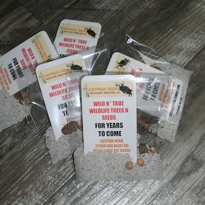 Puede incluir: Primer plano de un paquete de semillas de árboles silvestres "Wild N' True" para años venideros. El paquete es blanco con texto negro y un logotipo de ciervo rojo. El paquete está sobre una superficie de madera.