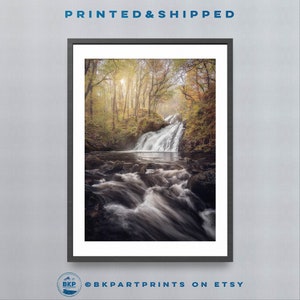May include: A photograph of a waterfall in a forest. The water is flowing over rocks and into a pool below. The trees are green and yellow, and the sun is shining through the leaves.