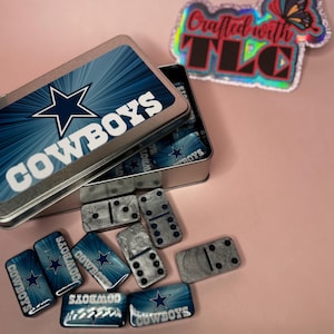 May include: A domino set with a blue and white Cowboys theme. The dominoes are silver with black dots. The set comes in a metal tin with the Cowboys logo and the word "Cowboys". A holographic sticker with the text "Crafted with TLC" is also visible.