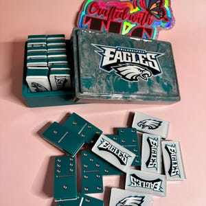 May include: A teal and white domino set featuring the Philadelphia Eagles logo. The set includes a storage box and individual dominoes with white and teal accents. A holographic sticker with the text "Crafted with Love" is also visible.