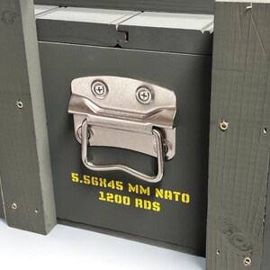 K&ouml;nnte beinhalten: Eine olivgr&uuml;ne Holzkiste mit einem silbernen Metallgriff und Verschlussmechanismus. Auf der Vorderseite befindet sich gelber Schablonentext mit der Aufschrift 5.56X45 MM NATO 1200 RDS. Die Kiste ist aus vertikalen Holzlatten mit sichtbaren Schrauben.