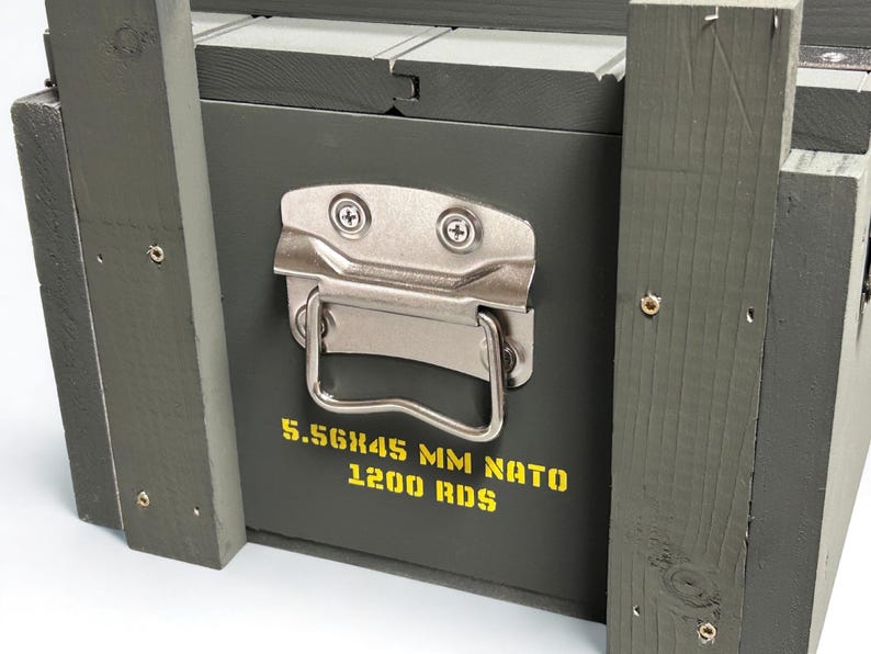K&ouml;nnte beinhalten: Eine olivgr&uuml;ne Holzkiste mit einem silbernen Metallgriff und Verschlussmechanismus. Auf der Vorderseite befindet sich gelber Schablonentext mit der Aufschrift 5.56X45 MM NATO 1200 RDS. Die Kiste ist aus vertikalen Holzlatten mit sichtbaren Schrauben.