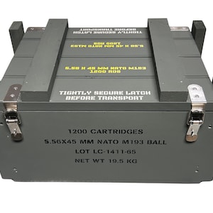 K&ouml;nnte beinhalten: Eine rechteckige graue Holzkiste mit Metallverschl&uuml;ssen. Auf der Oberseite steht in gelber Schrift TIGHTLY SECURE LATCH BEFORE TRANSPORT und 5.56 X 45 MM NATO M193 1200 RDS. Die Vorderseite zeigt schwarzen Text: 1200 CARTRIDGES, 5.56X45 MM NATO M193 BALL, LOT LC-1411-65, NET WT 19.5 KG.