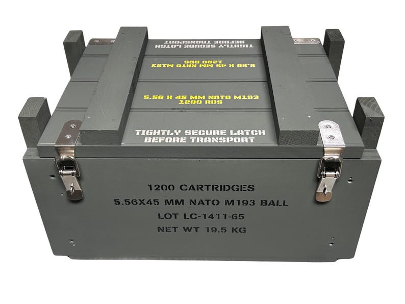 K&ouml;nnte beinhalten: Eine rechteckige graue Holzkiste mit Metallverschl&uuml;ssen. Auf der Oberseite steht in gelber Schrift TIGHTLY SECURE LATCH BEFORE TRANSPORT und 5.56 X 45 MM NATO M193 1200 RDS. Die Vorderseite zeigt schwarzen Text: 1200 CARTRIDGES, 5.56X45 MM NATO M193 BALL, LOT LC-1411-65, NET WT 19.5 KG.