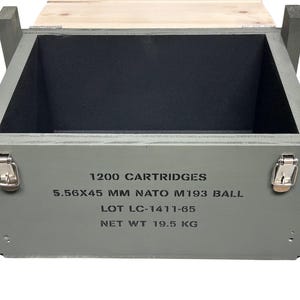 K&ouml;nnte beinhalten: Eine offene rechteckige Holzkiste in Olivgr&uuml;n mit schwarzem Innenraum. Die Vorderseite zeigt schwarzen Schablonentext: 1200 CARTRIDGES, 5.56X45 MM NATO M193 BALL, LOT LC-1411-65, NET WT 19.5 KG. Sie verf&uuml;gt &uuml;ber zwei silberne Metallverschl&uuml;sse und einen Deckel.