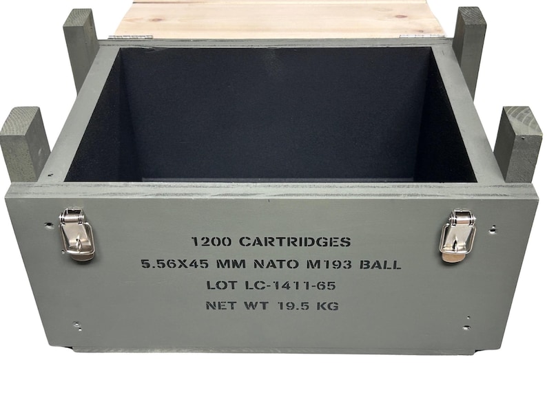 K&ouml;nnte beinhalten: Eine offene rechteckige Holzkiste in Olivgr&uuml;n mit schwarzem Innenraum. Die Vorderseite zeigt schwarzen Schablonentext: 1200 CARTRIDGES, 5.56X45 MM NATO M193 BALL, LOT LC-1411-65, NET WT 19.5 KG. Sie verf&uuml;gt &uuml;ber zwei silberne Metallverschl&uuml;sse und einen Deckel.