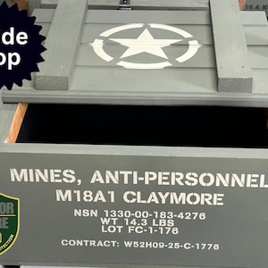 May include: A gray wooden box with a sliding top, featuring a white star emblem. The box is labeled "MINES, ANTI-PERSONNEL M18A1 CLAYMORE" and weighs 14.3 lbs. The box has a "Slide Top" label.