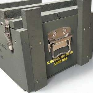 K&ouml;nnte beinhalten: Eine olivgr&uuml;ne Munitionskiste aus Holz mit silbernen Metallverschl&uuml;ssen und Griffen. An der Seite befindet sich der gelbe Aufdruck 5.56X45 MM NATO 1200 RDS. Der Deckel ist mit einem Metallverschluss gesichert und die Kiste zeigt ein Nettogewicht von 19,5 KG.