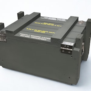 K&ouml;nnte beinhalten: Eine olivgr&uuml;ne, rechteckige Munitionskiste aus Holz mit Metallscharnieren und verst&auml;rkten Ecken. Auf der Oberseite befindet sich gelber Schablonentext 5.56 X 45 MM NATO M193 1200 RDS und TIGHTLY SECURE LATCH BEFORE TRANSPORT in wei&szlig;er Schrift.
