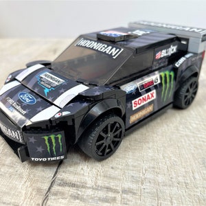 May include: Black and white Ford Fiesta rally car with Monster Energy and Toyo Tires logos. The car has a black and white American flag design on the hood and a clear windshield. The car is a Lego model.